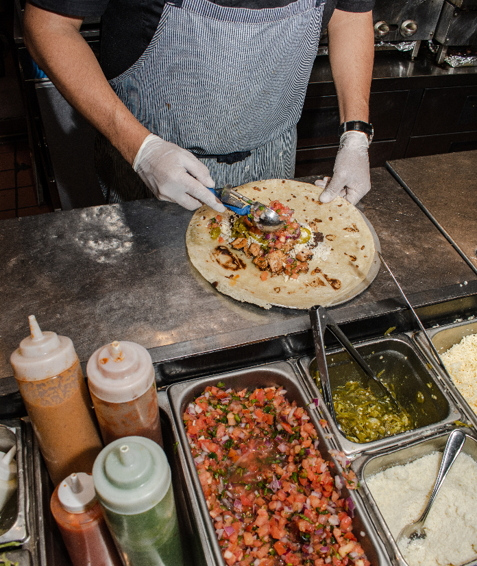 A cook assembling a taco on a flour tortilla at a kitchen prep station with pico de gallo, salsa verde, and sour cream in trays nearby.