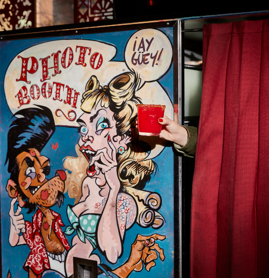 A colorful illustrated photo booth with cartoon characters and "Photo Booth" signage, with a hand holding a red cup emerging from the curtain.