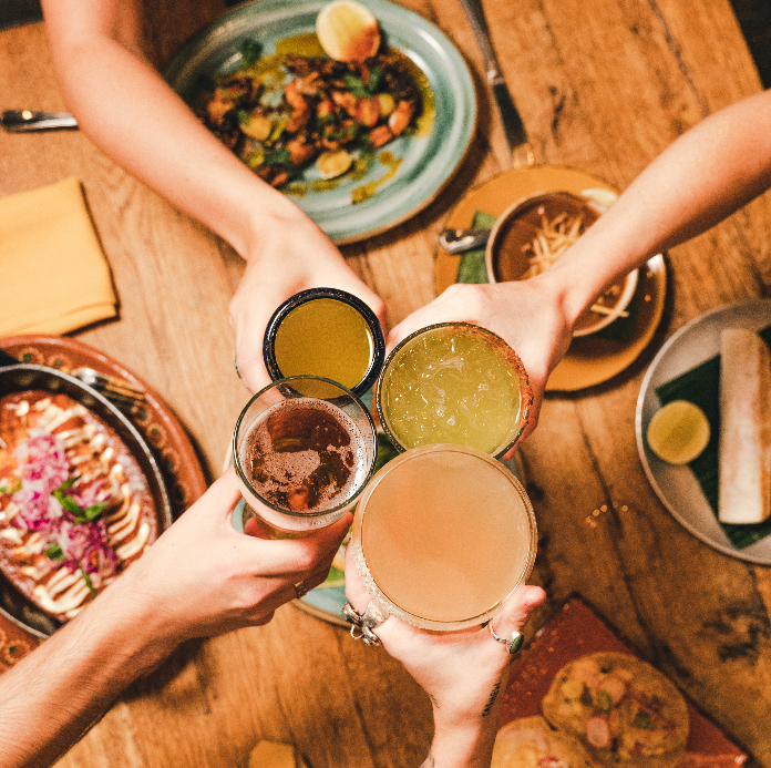 An overhead view of four hands toasting with drinks over a wooden table spread with Mexican dishes.