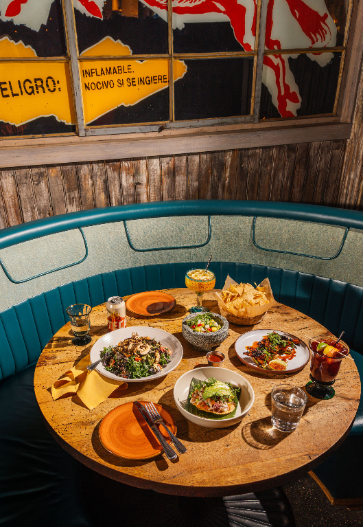 A round wooden table in a teal booth set with several Mexican dishes, chips, and drinks beneath vintage signage.