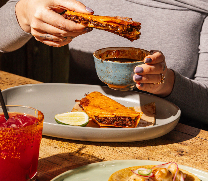 A person dipping a crispy taco into a bowl of broth, served with lime and a red drink on a wooden table.