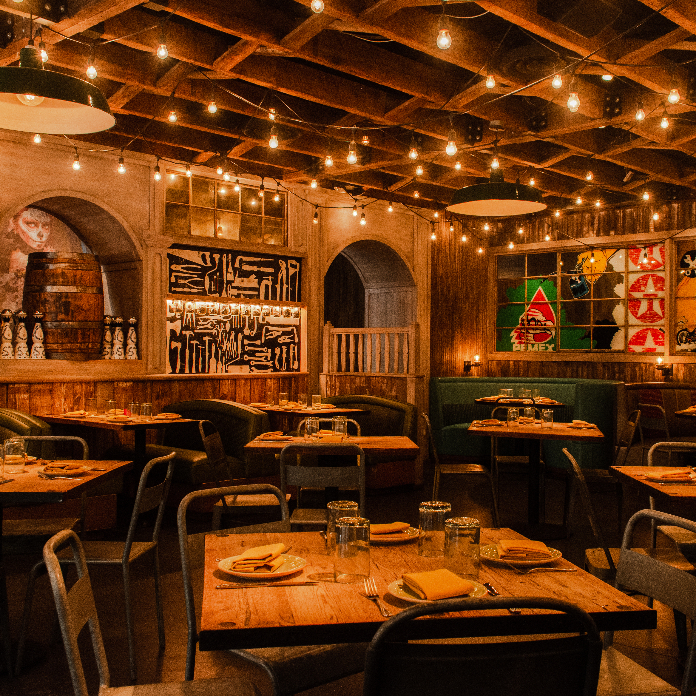 A warmly lit rustic dining room with wooden tables, string lights, brick arches, and colorful artwork on the walls.