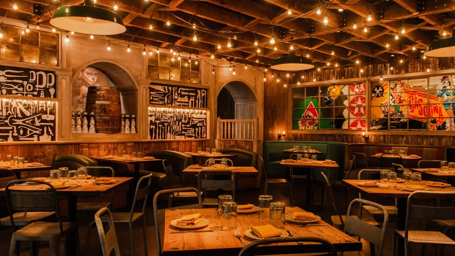 A warmly lit restaurant interior with wooden tables, industrial chairs, string lights, and bold graphic murals on the walls.
