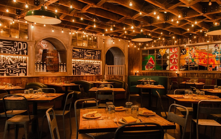 A warmly lit restaurant interior with wooden tables, industrial chairs, string lights, and bold graphic murals on the walls.