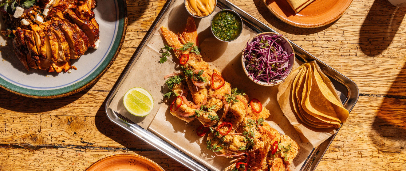 A metal tray with fried shrimp skewers, lime, salsa verde, red cabbage slaw, and tortilla chips on a wooden table.